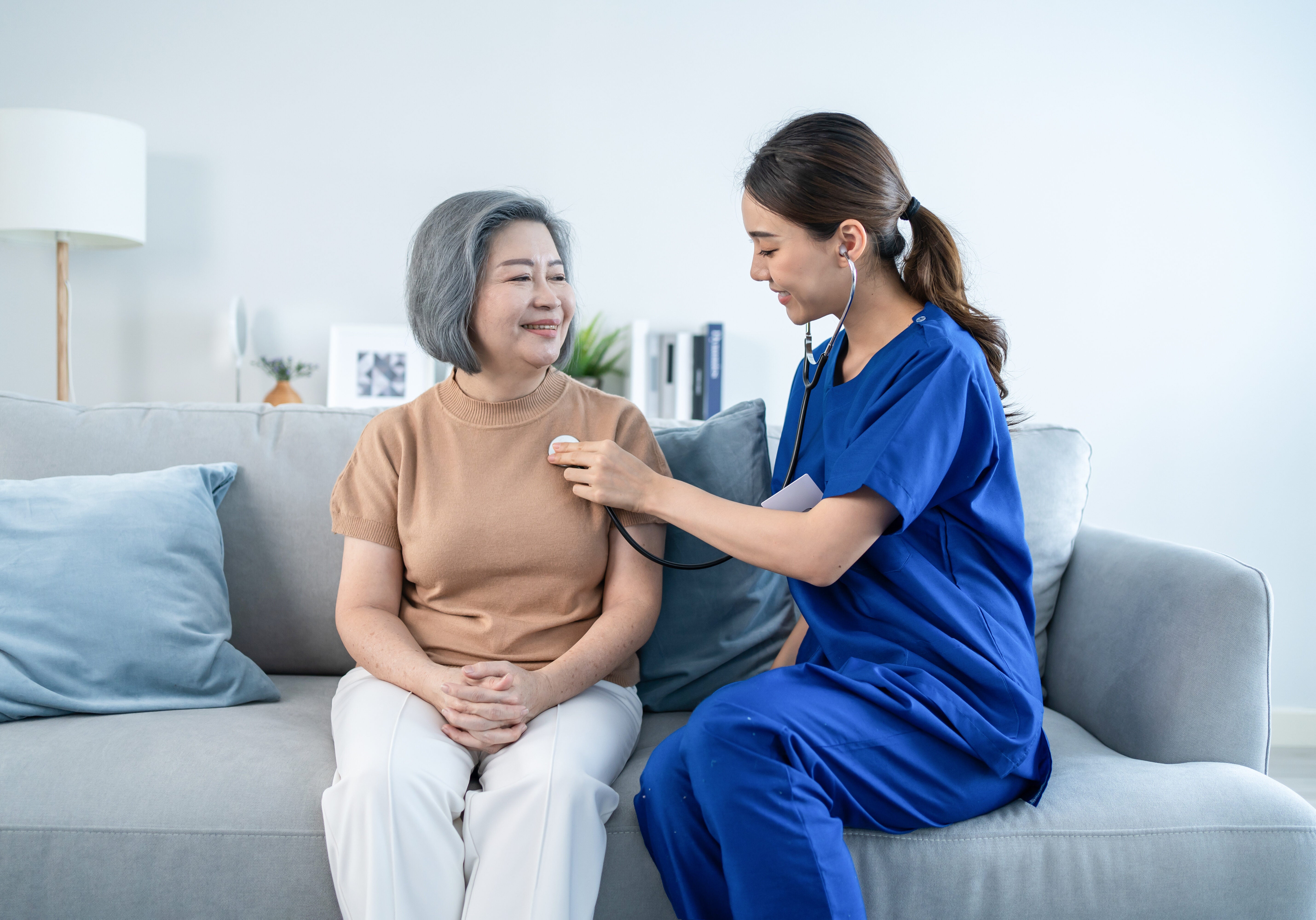 Asian attractive nurse at nursing home take care of senior elder woman. Beautiful therapist doctor measure heart rate by stethoscope on female older patient on sofa. Medical insurance service concept.