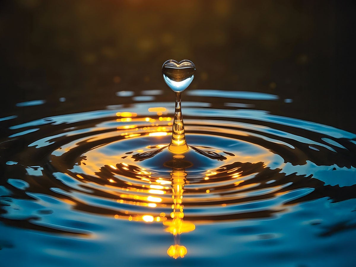 A water droplet forming a heart shape, with ripples on the water reflecting the sunset.