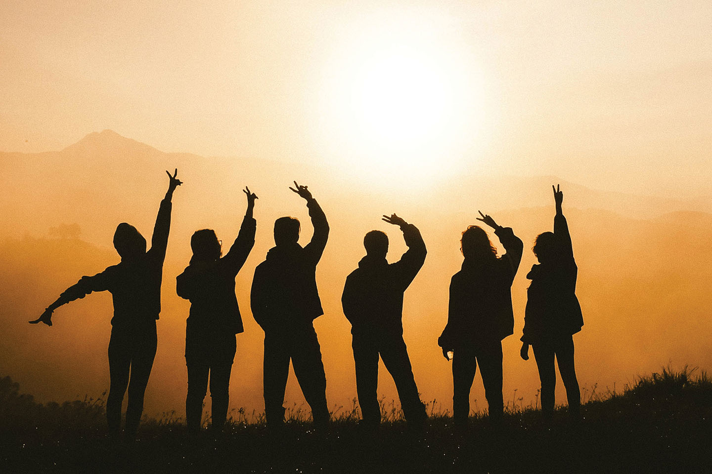 A landscape photo of several youth, with their silhouettes standing atop of a grassy hill amdist a sunset.