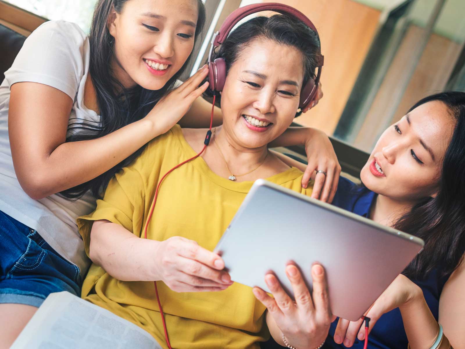 Stock photo of two Asian ladies helping an Asian aunt use an iPad wearing earphones.