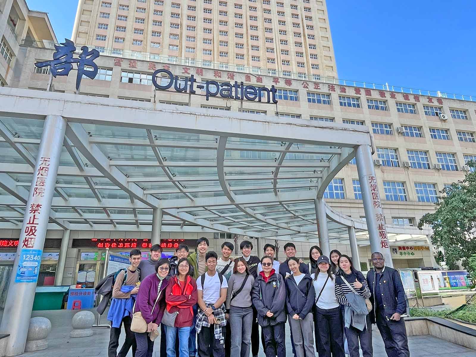 Various Chinese Asians standing outside a hospital entrance that has a sign on-top reading Outpatient.