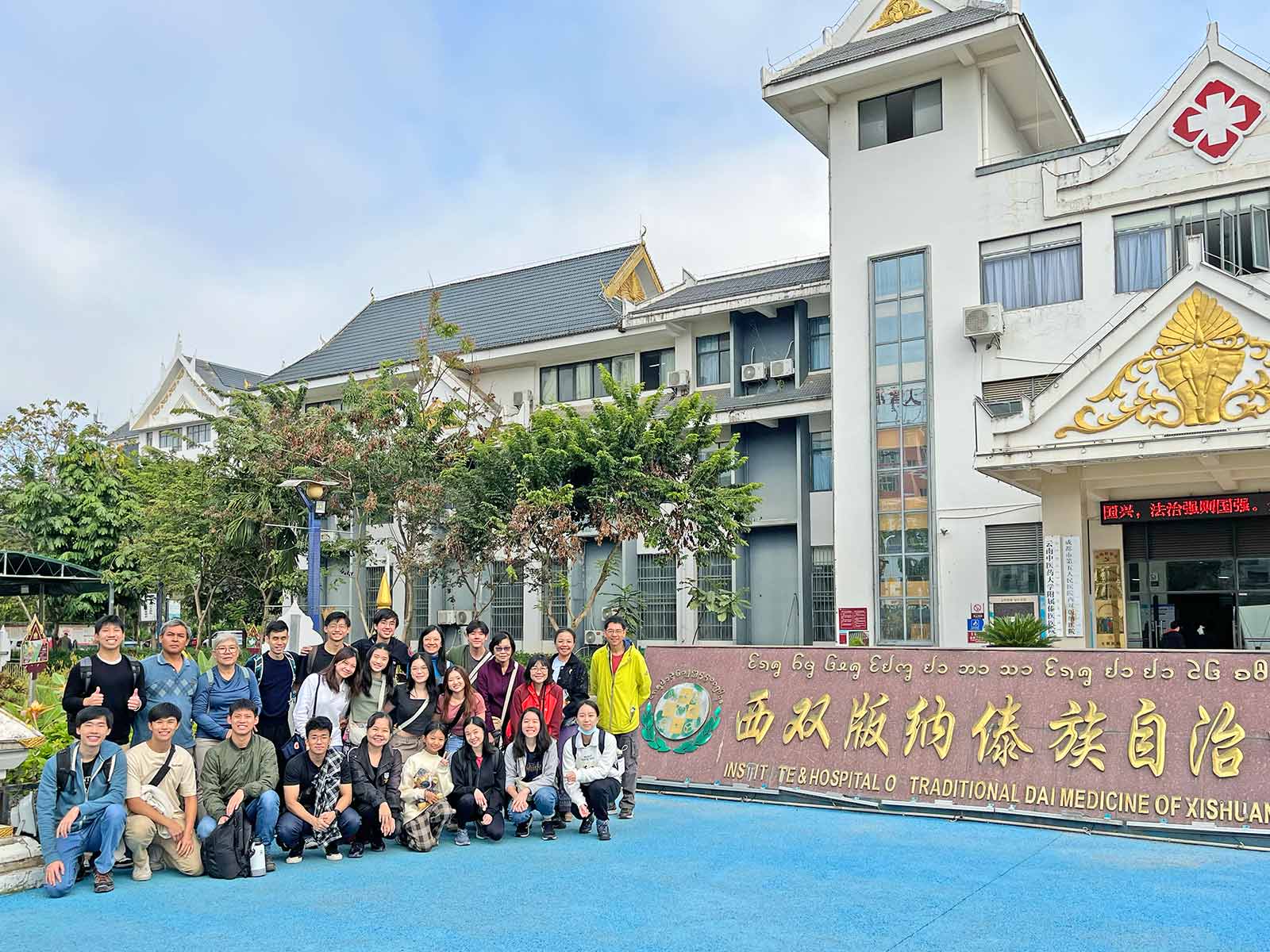 Various students, staff, team members standing to the left of Traditional Dai Medicine Hospital.