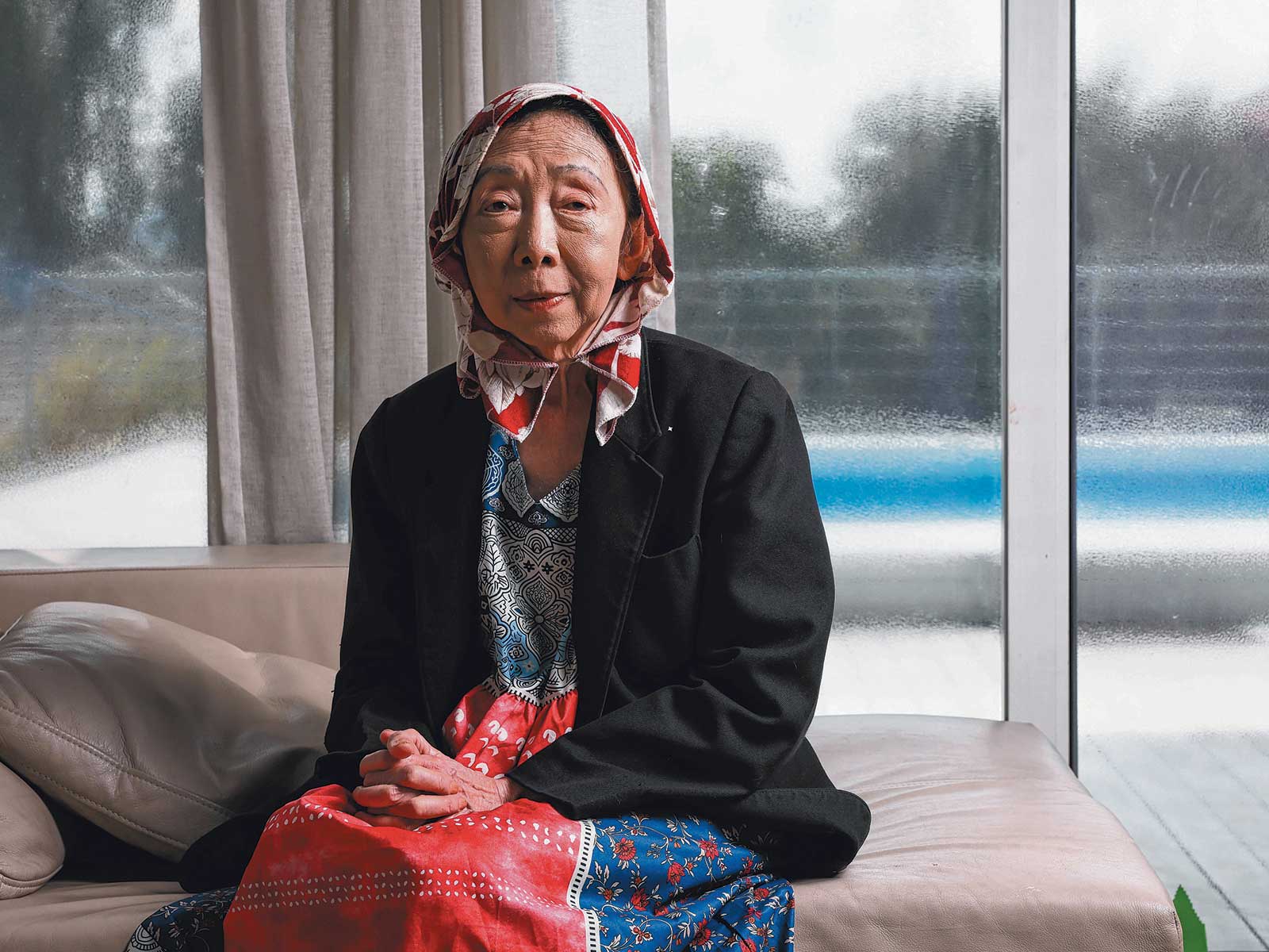 Portrait photo of an elderly woman wearing a red and white scarf, black blazers, and a red and blue dress with floral patterns sitting on a pale-brown sofa.