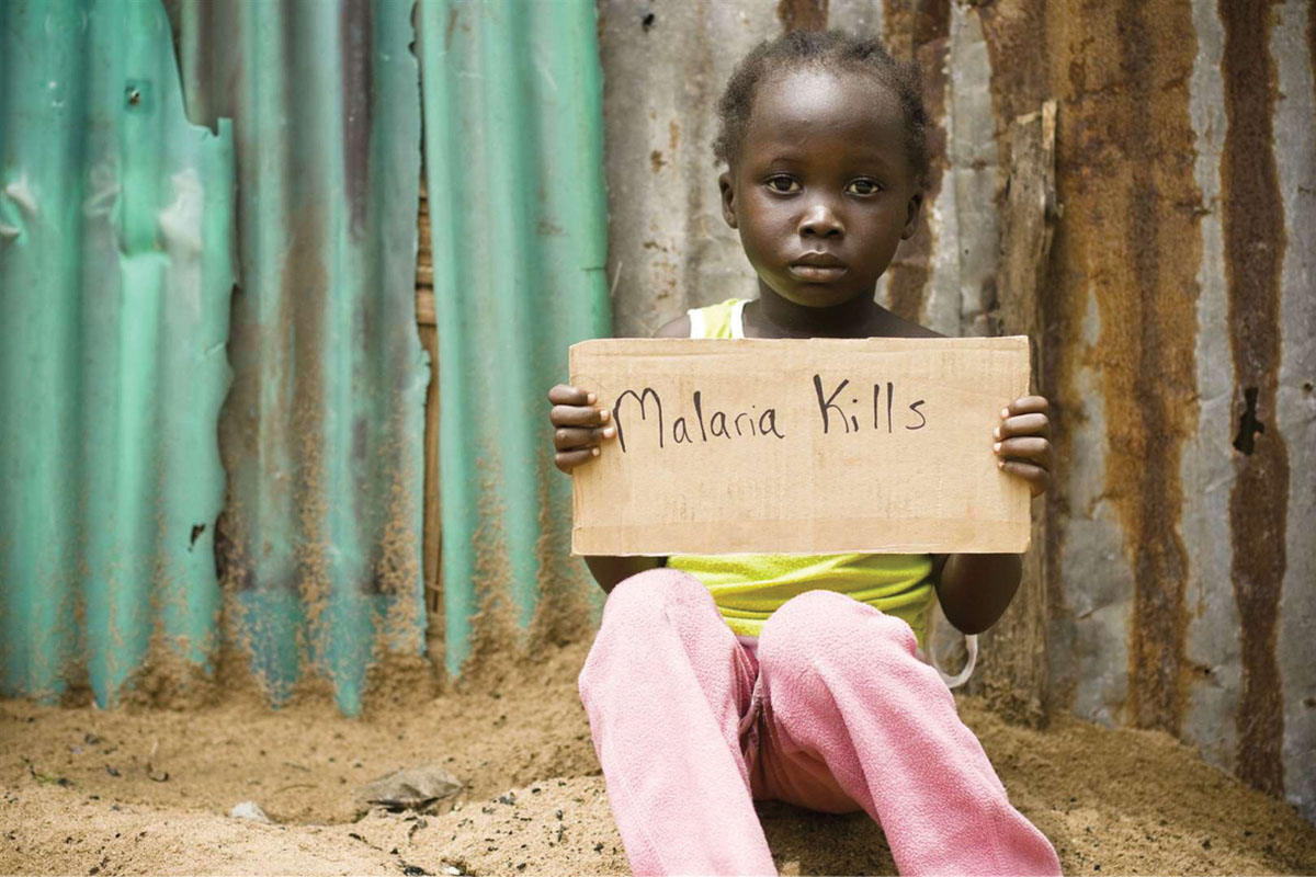 Photo of a dark-skinned young person sitting on the floor, carrying the sign that reads Malaria Kills.