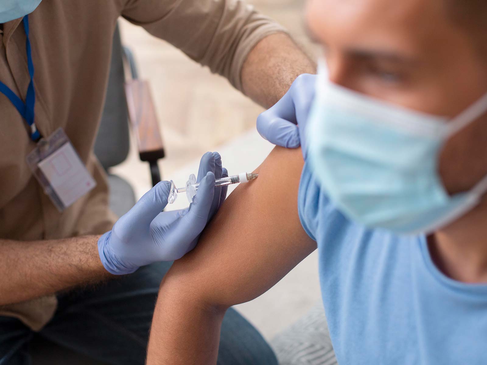 Photo of dark-skinned person wearing mask receiving injection from doctor in brown shirt