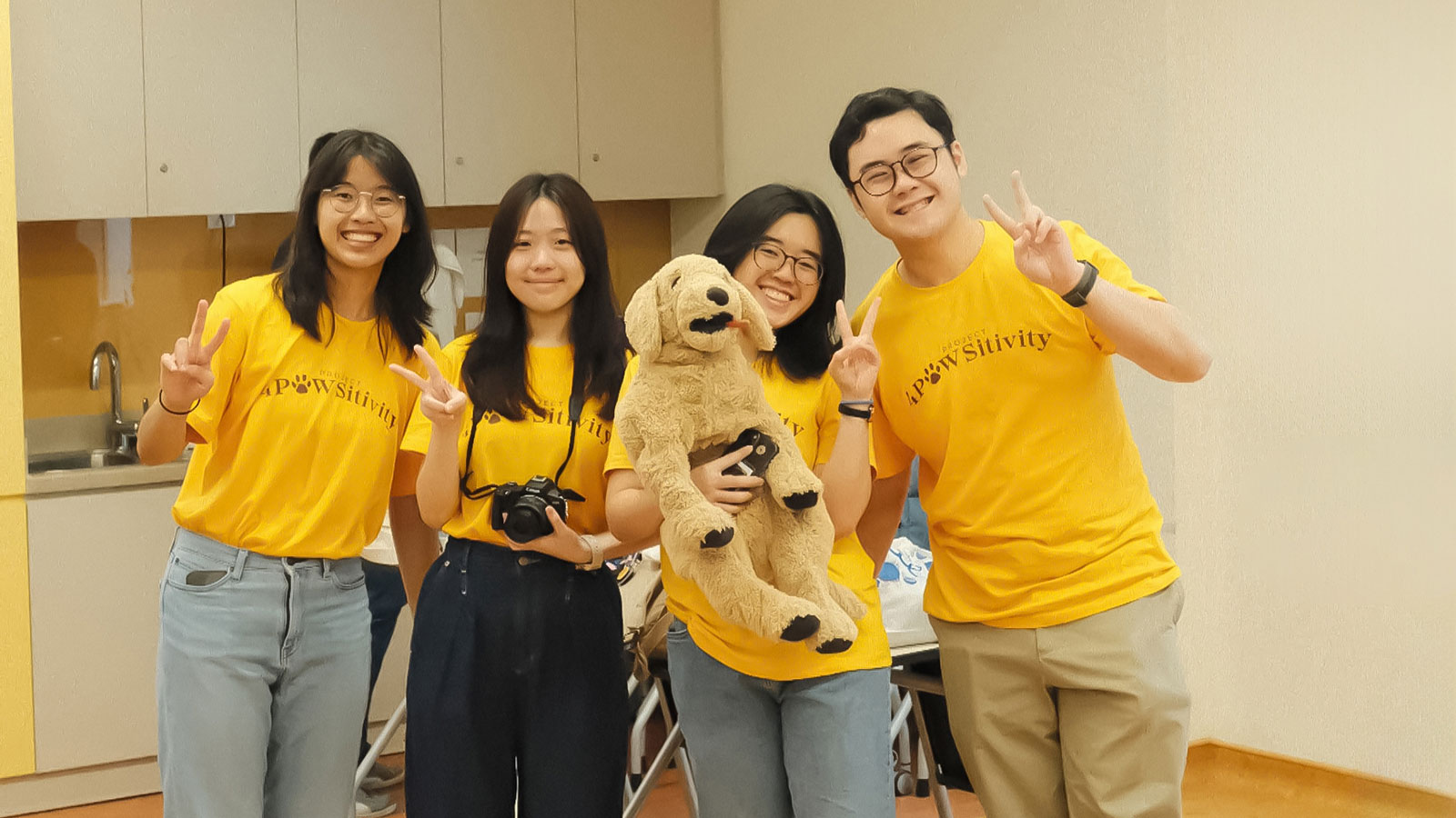 Group photo of 3 ladies and 1 guy wearing a yellow shirt that reads 4Pawsitivity. 3rd girl from left is carrying a plush dog.