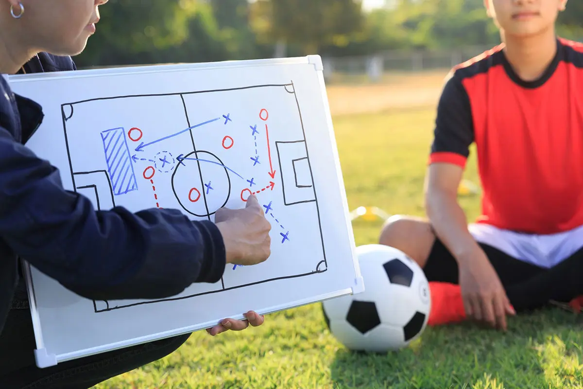 Two football players seated on the grass, the left player using a clipboard to explain tactics to the right player.