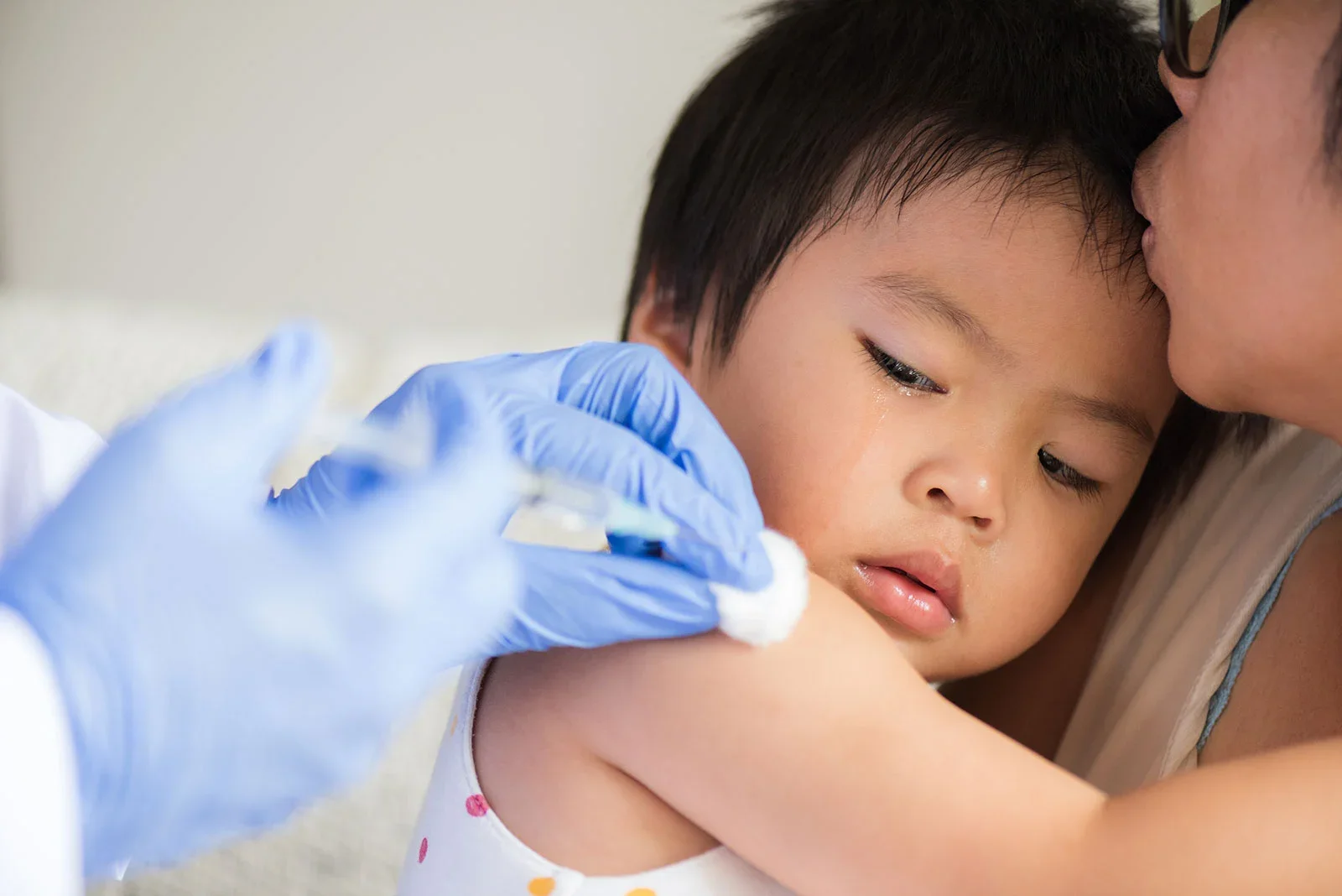 A doctor wearing blue gloves administering a measels vaccine shot to a short-haired baby carried by a bespetacled mother.