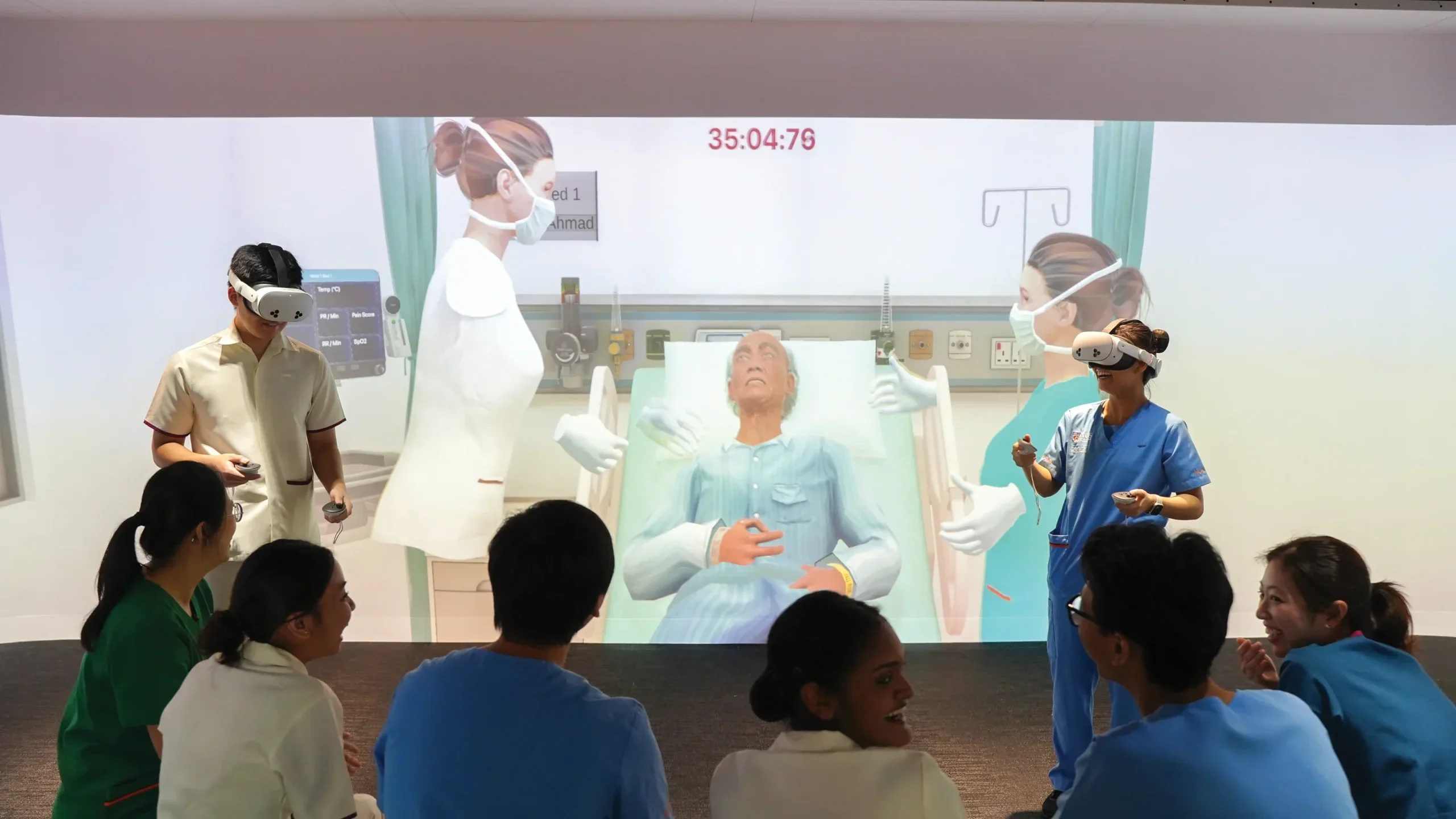 6 trainee nurses look at 2 other trainee nurses on VR headsets, with the projection screen at the middle of the room showing a simulation of nurses performing care duties onto a virtual patient.