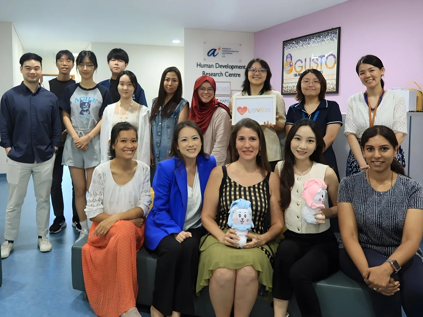 A group photo of 15 smiling people gathered in the Human Development Research Centre. Five women are seated in the front row, two holding small plush toys, while ten people stand behind them, one holding a framed sign featuring a heart and the word LOVING.