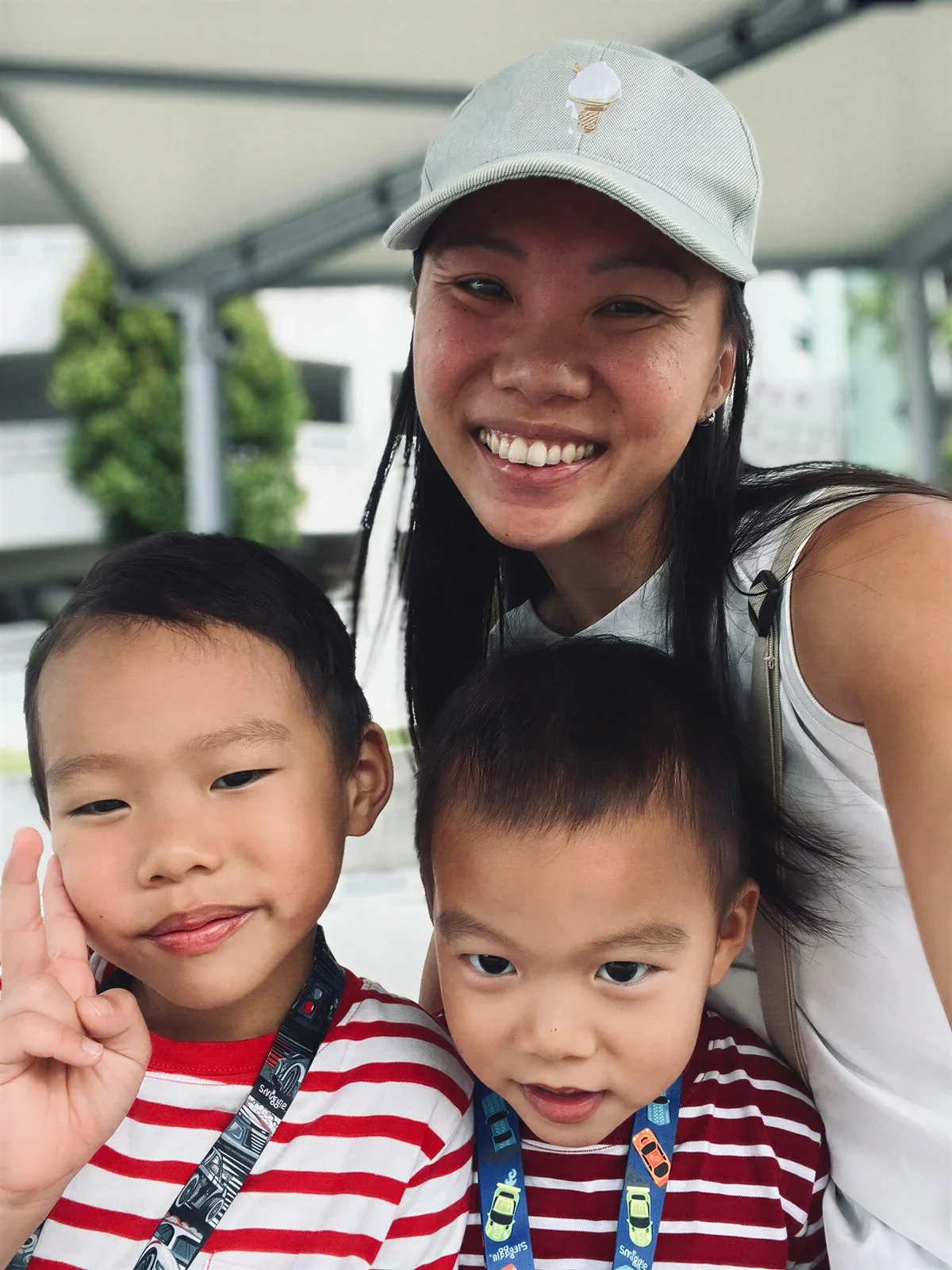 A smiling woman taking a selfie outdoors with two young boys. The boy on the left is holding up a peace sign, and both boys are wearing matching red and white striped shirts with colorful lanyards.