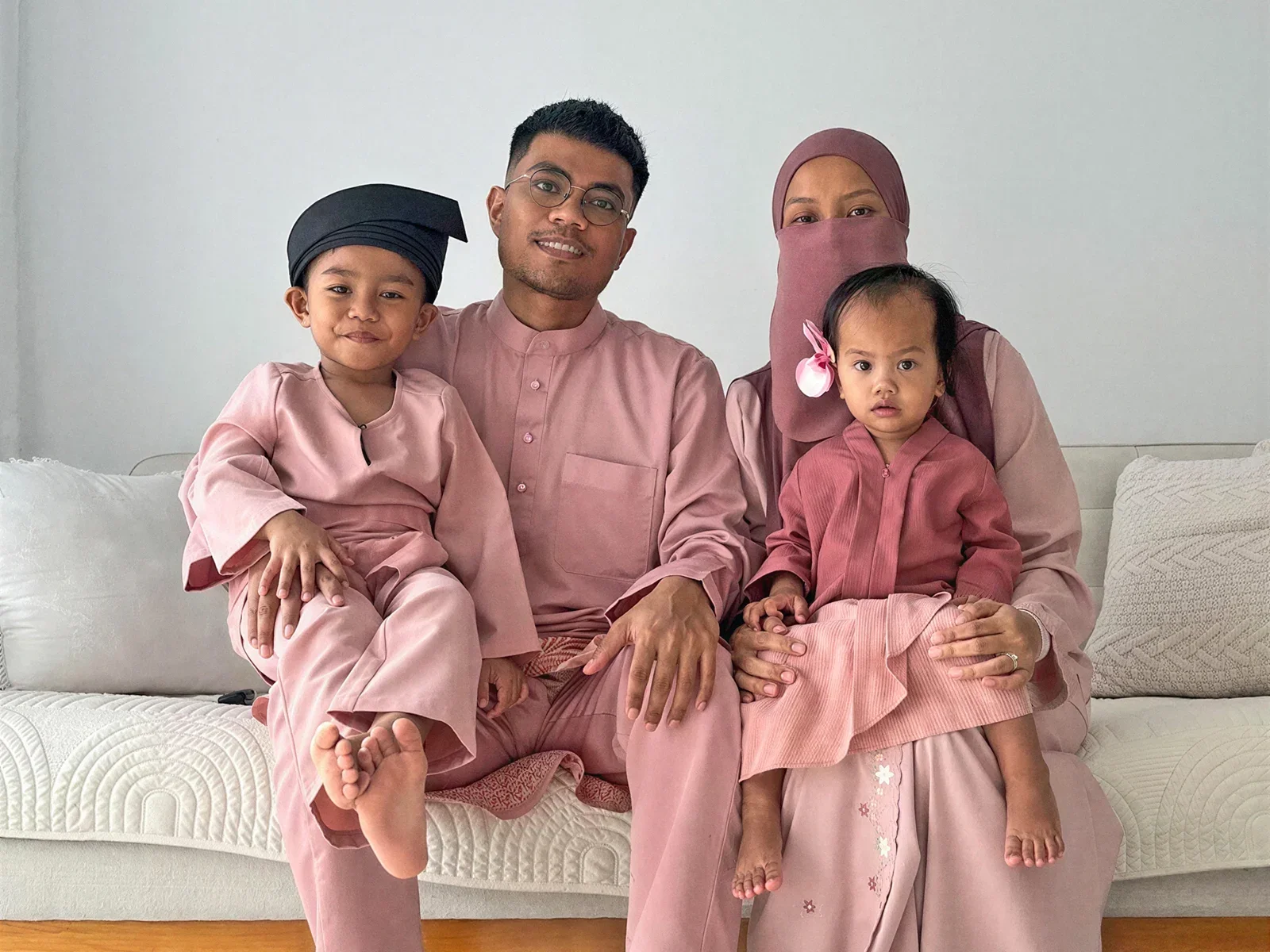 A family of four sitting together on a white sofa, dressed in beautifully coordinated pink traditional Malay attire. The smiling father holds their young son, who wears a black traditional headpiece, while the mother, wearing a matching pink hijab and niqab, holds their toddler daughter on her lap.