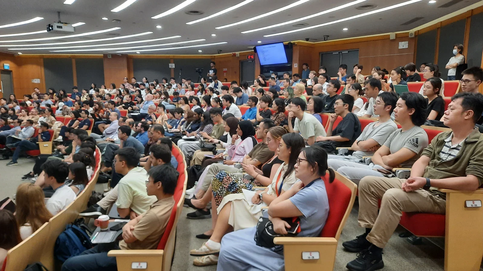 A brightly-lit lecture room full of attendees, with red cushion seats, orange-brown walls and gray flooring.