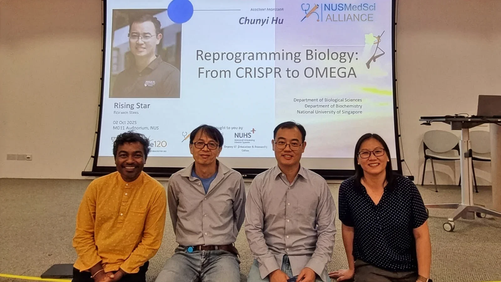 From left to right, a male Indian professor in an orange shirt and black pants, a male bespectacled Chinese professor in a gray long-sleeved shirt and a blue t-shirt underneath and blue pants, another male bespectacled Chinese professor in a gray long-sleeved shirt and blue jeans but tucked out, and a female bespectacled Chinese professor in dark blue short-sleeve shirt and khaki-brown pants.