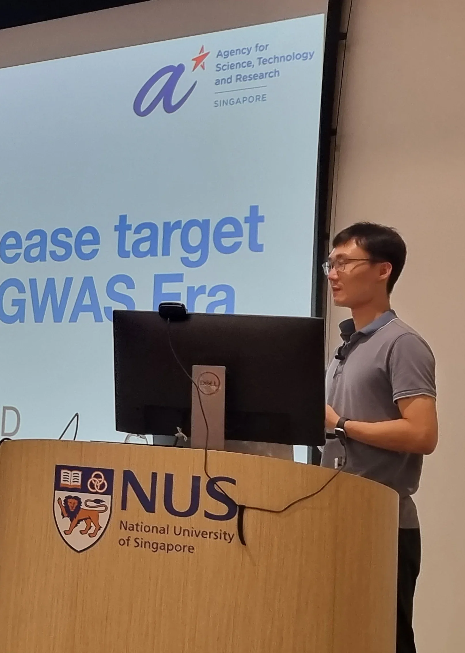A bespectacled male Chinese professor in a gray short-sleeve polo tee gives a speech at the podium with a NUS logo in front of it. The presentation slide reads, Identification of disease target genes in the Post-GWAS Era.