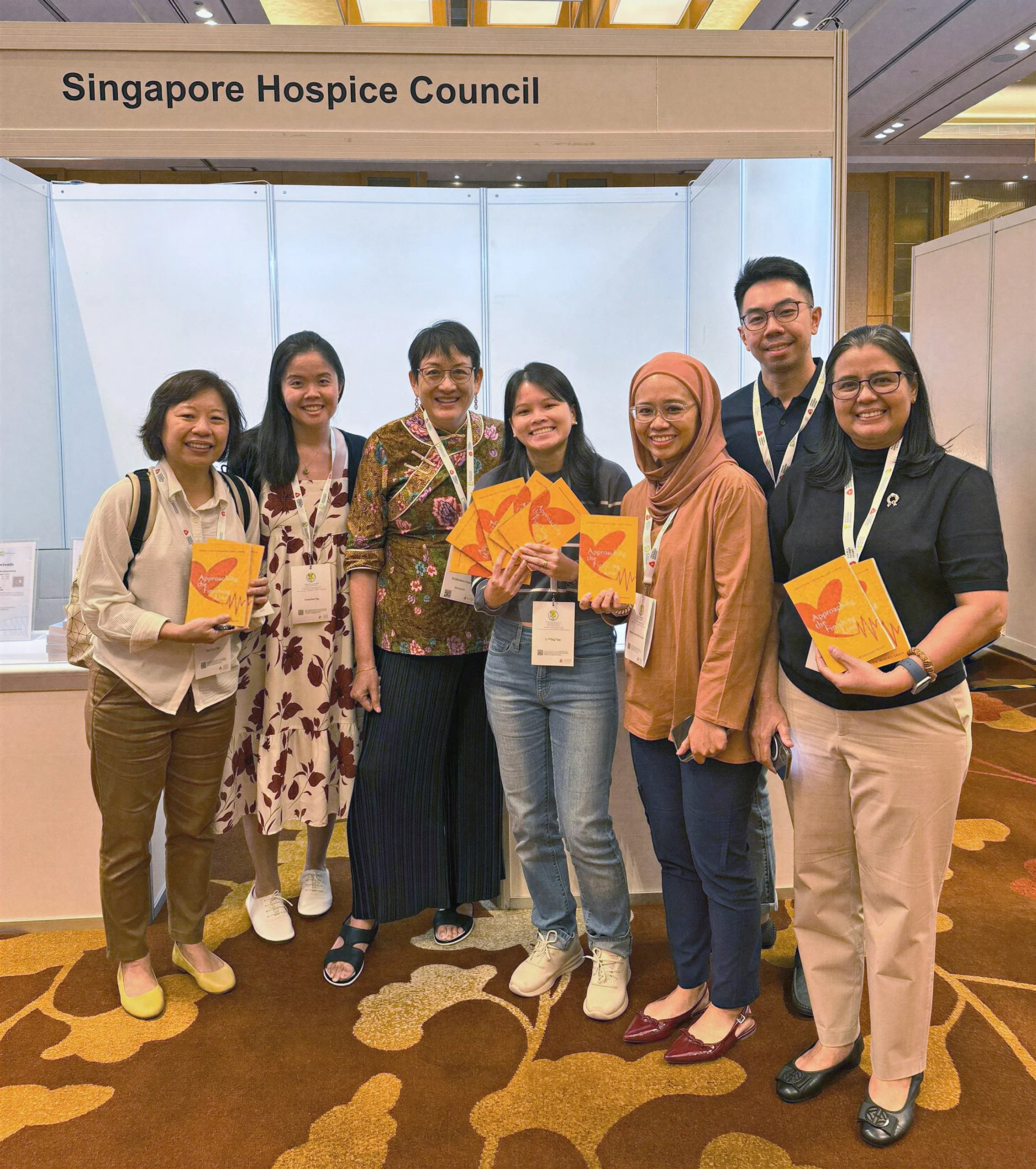 Doctor Noreen Chan, bespectacled and having a lanyard, and 6 other persons outside the Singapore Hospice Council booth.